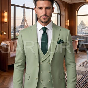 Peut inclure: Un costume trois pièces vert clair, comprenant une veste, un gilet et un pantalon, associé à une chemise blanche et une cravate vert foncé. Une pochette vert foncé est visible. En arrière-plan, une ville avec une tour.