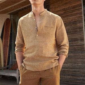 May include: A man wearing a tan linen shirt with a mandarin collar and matching shorts. The shirt has a single chest pocket and button details. He is wearing sunglasses. The background includes surfboards and a wooden structure.