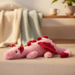 May include: A soft pink dragon toy with red wings, ear tips, and dorsal spikes. The plush dragon is lying down on a neutral surface. The toy has a friendly expression and is designed to be cuddly. The dragon is a children's toy.