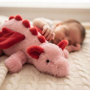 May include: A pink plush dragon toy with red wings and spikes lies on a cream-coloured blanket. The toy has black eyes and is positioned near a sleeping baby, creating a gentle scene.