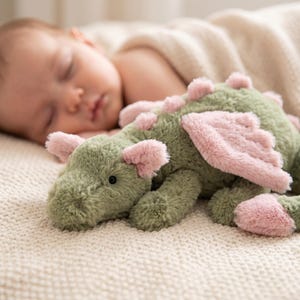May include: A plush green dragon toy with pink accents, including wings and spikes, rests on a cream-colored blanket. The soft toy has a friendly expression with a black eye. The dragon is a children's toy.