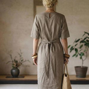 Puede incluir: Vestido de lino marrón claro con mangas cortas y cintura anudada. El vestido se combina con sandalias marrones y un bolso tote tejido. La modelo lleva pulseras de madera. El vestido es de largo midi.