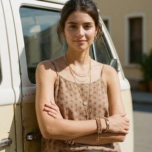 May include: A woman wearing gold jewelry, including layered necklaces, hoop earrings, and bracelets. She is wearing a patterned tank top with a brown and red design. The background includes a vintage van and a building.