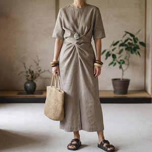May include: A beige linen dress with short sleeves and a knotted waist, worn with a woven tote bag, wooden bracelets, a pendant necklace, and brown sandals. The dress is a neutral colour. The overall style is minimalist and natural, with a focus on texture.