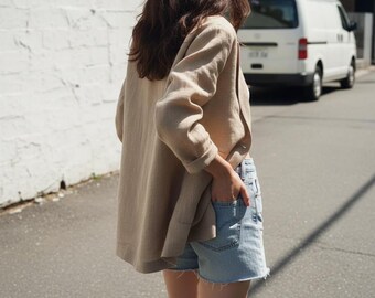 May include: A tan linen blazer with rolled-up sleeves and light blue denim shorts. The blazer is open, revealing the shorts and a hand in the pocket. The outfit is casual and suitable for warm weather.