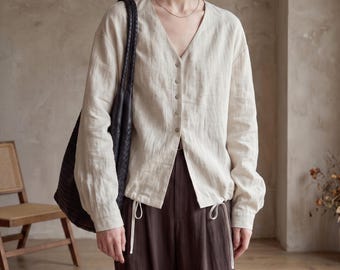 May include: A beige long-sleeved button-down shirt with a V-neckline, paired with dark brown trousers featuring white drawstring ties at the waist. A large black woven tote bag hangs over one shoulder, completing the ensemble.