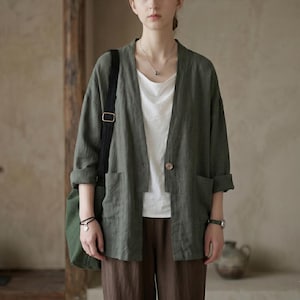 May include: An olive green linen jacket with a single button closure and two front pockets. The jacket is worn over a white top and paired with brown pants. A black shoulder bag and a watch are also visible.