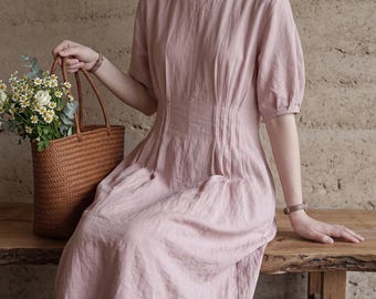 May include: A dusty pink midi dress with short puff sleeves, a round neckline, and vertical pleats at the waist. The dress features a textured fabric and a flowing skirt. Beside the dress is a brown woven tote bag filled with white daisies.