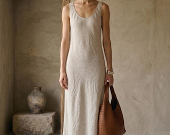 May include: A sleeveless, floor-length beige dress with a scoop neckline and a straight silhouette, paired with a large, woven brown leather hobo bag featuring a textured, interlaced pattern and a single shoulder strap, worn with a silver bracelet.
