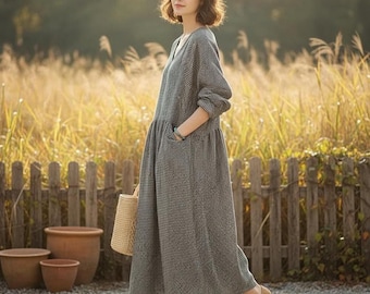 May include: A long-sleeved, gray and white checkered dress with a V-neck and pockets. The dress is paired with brown shoes and a straw bag. The background features a wooden fence, potted plants, and tall grass.