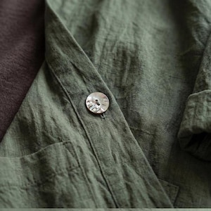 Pu&ograve; includere: Primo piano di una camicia di lino verde con un singolo bottone perlato. La camicia ha una vestibilit&agrave; rilassata, una tasca visibile e maniche arrotolate. Il tessuto ha un aspetto strutturato, suggerendo una fibra naturale. Il colore &egrave; un verde tenue e terroso.