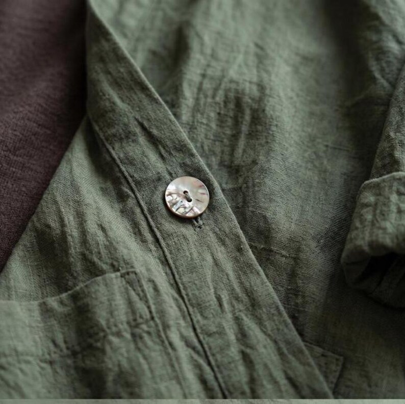 Pu&ograve; includere: Primo piano di una camicia di lino verde con un singolo bottone perlato. La camicia ha una vestibilit&agrave; rilassata, una tasca visibile e maniche arrotolate. Il tessuto ha un aspetto strutturato, suggerendo una fibra naturale. Il colore &egrave; un verde tenue e terroso.