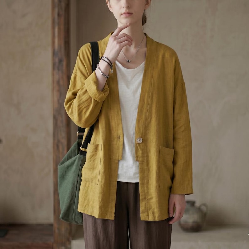 Pu&ograve; includere: Giacca in lino giallo senape con un solo bottone e due tasche anteriori. La giacca &egrave; indossata sopra un top bianco e abbinata a pantaloni marroni. Una borsa tote verde &egrave; portata a tracolla.