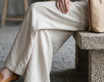 May include: Wide-leg trousers in a light beige, textured fabric with a loose, flowing fit. The trousers are paired with a tan leather-trimmed canvas tote bag and brown leather loafers, creating a neutral-toned ensemble.