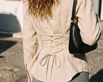May include: A beige long-sleeved button-down shirt featuring a corset-style lace-up back with matching fabric ties. The shirt is paired with a black shoulder bag and a gray skirt, showcasing a fitted silhouette through the back lacing detail.