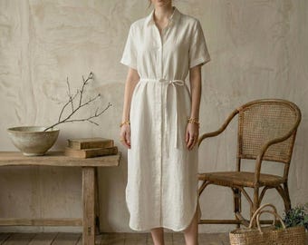 May include: A woman wearing a white linen shirt dress with short sleeves and a matching belt. The dress is buttoned down the front and falls to mid-calf. She is wearing brown sandals. The background includes a wooden bench, a wicker chair, and a woven basket.