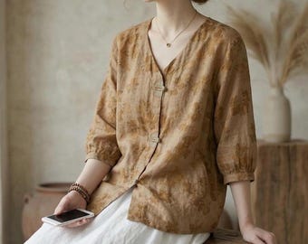 Può includere: Una camicetta beige con motivo floreale, scollo a V e maniche a tre quarti. La camicetta è abbottonata e indossata con una gonna bianca. Il tessuto sembra essere lino o un materiale leggero simile, ideale per il clima caldo.