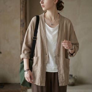 Pu&ograve; includere: Giacca di lino beige con due tasche frontali e una chiusura con un solo bottone. La giacca &egrave; indossata sopra un top bianco e abbinata a pantaloni marroni. Una borsa di tela verde con una tracolla nera &egrave; portata a spalla.
