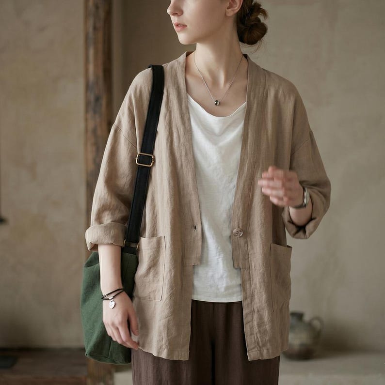 Pu&ograve; includere: Giacca di lino beige con due tasche frontali e una chiusura con un solo bottone. La giacca &egrave; indossata sopra un top bianco e abbinata a pantaloni marroni. Una borsa di tela verde con una tracolla nera &egrave; portata a spalla.