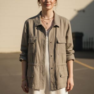 Puede incluir: Chaqueta utilitaria marrón claro con cuatro bolsillos delanteros y cintura con cordón. La chaqueta está abotonada y se usa sobre una blusa blanca. La modelo también lleva collares y pulseras doradas.