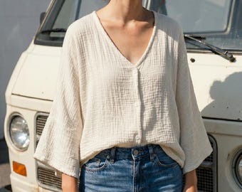 May include: Cream-colored, long-sleeve button-down shirt with a V-neck and wide sleeves. The shirt is made of a textured fabric and is paired with blue jeans. The background features a vintage white van.