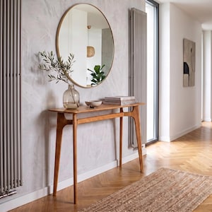Op de afbeelding: Een lichtbruine houten sidetable met vier taps toelopende poten en een rechthoekig blad. Op de tafel staat een doorzichtige glazen vaas met een groene tak, een kleine keramische kom en een stapel boeken. Een ronde spiegel hangt aan de muur.
