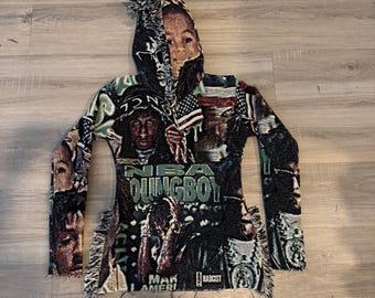 May include: A woven hoodie with a collage design, including a portrait and the text "NBA YOUNGBOY". The hoodie has a fringed hem and a hood. The colour scheme includes black, green, and tan.