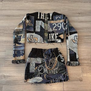 May include: A two-piece set featuring a cropped jacket and a mini skirt. The set is made from a tapestry fabric with a black, blue, and beige color scheme. The jacket has long sleeves and the skirt features a panther graphic with the text "PVAMU".