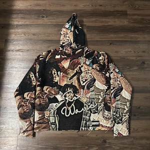 May include: A tapestry hoodie featuring a collage of faces and the text "Road Wave" in white script. The hoodie is predominantly black, brown, and beige, with a raw hemline. The hood is patterned with faces.