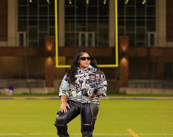 May include: A person wearing sunglasses, a patterned shirt, and black leather pants stands on a football field. The person is holding a camera and has one foot on a purple and white ball. The background features a stadium with the text "PRAIRIE VIEW A&M UNIVERSITY".