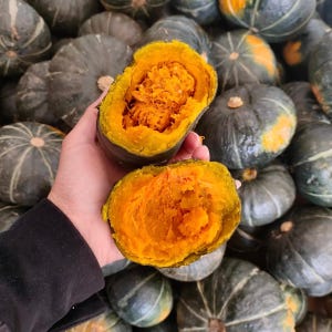 Puede incluir: Una calabaza cocida cortada por la mitad, con pulpa de color naranja brillante, se sostiene frente a una pila de calabazas enteras. El interior de la calabaza es de color naranja vibrante, contrastando con el exterior verde oscuro. El fondo presenta muchas calabazas enteras.