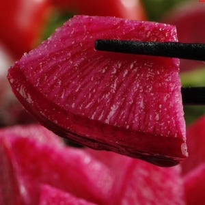 May include: A close-up of a vibrant, magenta-colored radish slice held by black chopsticks. The radish has a textured surface and appears juicy. Other radish slices are visible in the blurred background, suggesting a culinary theme.
