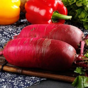 May include: Two elongated, deep red beets with textured skin are displayed on a wooden surface, alongside red and yellow bell peppers and leafy greens. The beets are the focal point, showcasing their vibrant color and shape, suggesting fresh produce.