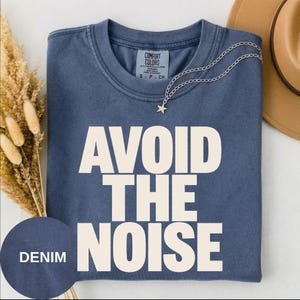 Può includere: Una t-shirt blu denim a girocollo con il testo bianco AVOID THE NOISE stampato in un carattere sans-serif in grassetto. La maglietta presenta un'etichetta Comfort Colors all'interno del collo ed è mostrata con una collana con ciondolo a stella.