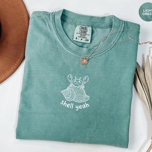 Puede incluir: Camiseta verde claro con un gráfico blanco de un cangrejo en una concha y la frase "shell yeah". Un collar dorado está sobre la camiseta. La camiseta está doblada sobre una superficie.