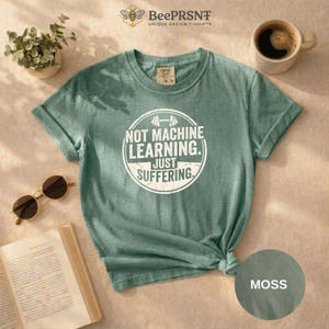 Puede incluir: Una camiseta verde salvia con el texto "NOT MACHINE LEARNING. JUST SUFFERING" en blanco. La camiseta tiene cuello redondo y mangas cortas. También se ven unas gafas de sol y una taza de café.
