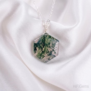 Puede incluir: Collar con colgante hexagonal de ágata musgosa. El colgante, con patrones verdes y blancos, está envuelto en alambre y suspendido de una cadena plateada. El collar descansa sobre una tela de satén blanca.