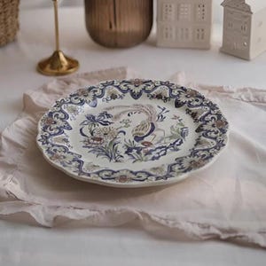 Peut inclure: Une assiette décorative en céramique blanc cassé avec un motif floral et d'oiseaux détaillé en bleu, marron et vert. L'assiette a un bord festonné et est posée sur un tissu rose.