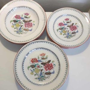 May include: A set of white ceramic plates with a floral and butterfly design. The plates feature a pink and red flower arrangement with yellow and blue accents. A brown and blue border surrounds the edge of the plates.