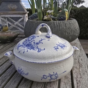 Puede incluir: Una fuente de cerámica blanca con tapa, adornada con motivos florales azules. La fuente tiene un asa curva en la tapa y asas laterales. La fuente está sobre una superficie de madera.