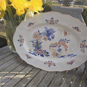 May include: An oval serving platter with a decorative floral and bird design in blue, orange, and brown. The white ceramic platter has a scalloped edge. A vase of yellow daffodils is next to the platter.