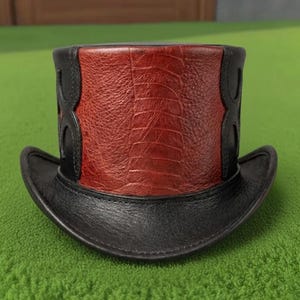 Può includere: Un cappello a cilindro in pelle nera e rossa. Il cappello presenta un pannello centrale rosso strutturato con ritagli decorativi neri e una tesa nera. Il cappello è su una superficie verde.