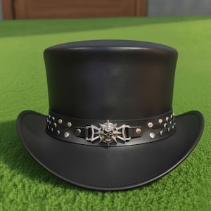 May include: A black top hat featuring a wide brim and a flat crown. A black leather-like band encircles the base, decorated with metallic silver studs and a central silver-toned emblem depicting a skull surrounded by a star-shaped cross design.