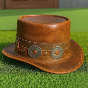 May include: A brown leather top hat featuring a wide band adorned with circular metallic medallions. Each medallion displays a central diamond-shaped inlay. The hat has a flat crown and a curved brim, showcasing a smooth, polished finish.