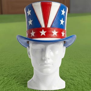 May include: A patriotic Uncle Sam hat with a blue brim and a red, white, and blue top. The hat features white stars and is displayed on a white mannequin head against a green background.