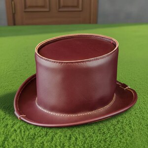 May include: A burgundy top hat with a wide brim. The hat is made of a smooth material and features a contrasting cream-colored stitch around the brim. The hat is sitting on a green surface.