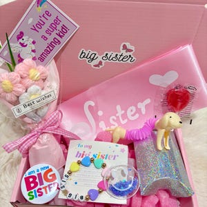 May include: A pink gift box containing a selection of gifts. Inside are a bouquet of pink flowers, a badge reading "BIG SISTER", a bracelet, and a pop tube toy. A card says "To my big sister".