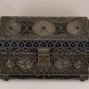 May include: Ornate rectangular jewelry box with a hinged lid. The box is silver-toned with intricate patterns, including circular and diamond shapes. Accents of red, yellow, and blue add color. The box has a clasp closure and small feet.