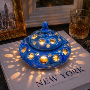 May include: A blue ceramic candle holder with a domed lid and circular cutouts, casting light patterns. The candle holder sits on a book titled "NEW YORK" with a photograph of the city. A framed picture and a glass are in the background.