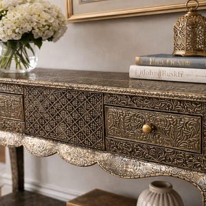Puede incluir: Consola ornamentada de estilo antiguo con tres cajones. La mesa presenta intrincados patrones florales y geométricos en un acabado dorado y bronce. Un jarrón de cristal con flores blancas, libros y una linterna decorativa encima.